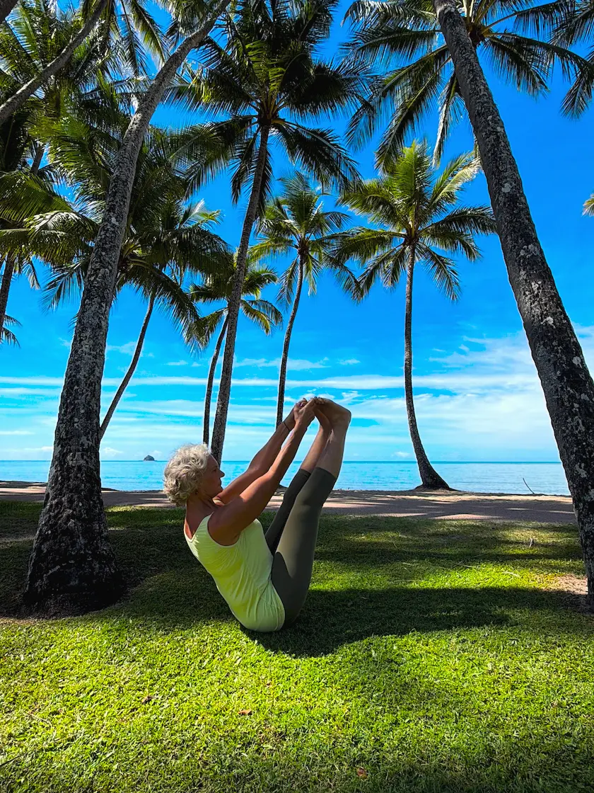 Susan Stackhouse seated on the grass under the palms at Kewarra Beach, holding both feet with her legs raised in Ubhaya Padangusthasana